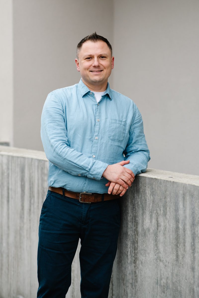Johannes Wachtel lächelt in die Kamera, im hellblauen Hemd an eine Mauer gelehnt